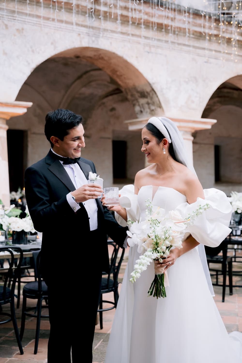 Bride and groom toast with cocktails beneath string-light arches at the colonial cloister reception.
