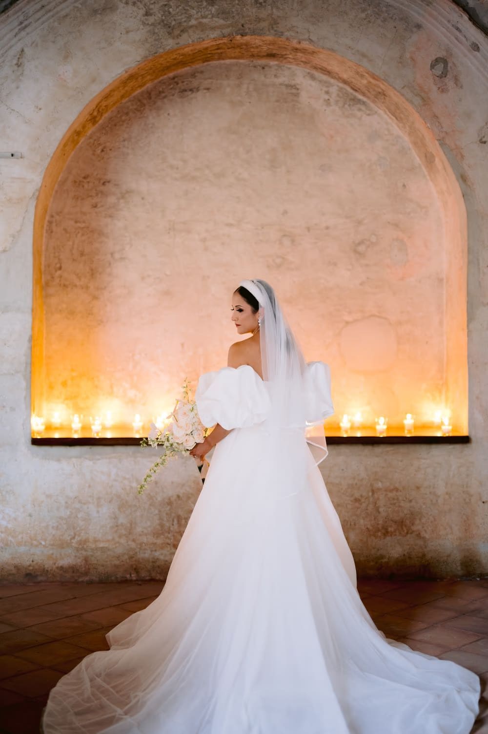Bride glances back in a warm candle-lit arched niche of Capuchinas Ruins — golden-hour bridal portrait.