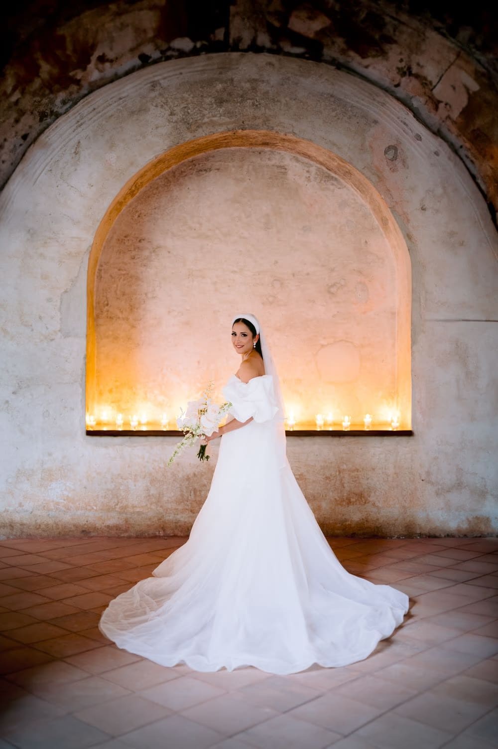 Wide bridal portrait — bride smiles holding her bouquet in front of an illuminated arched niche.