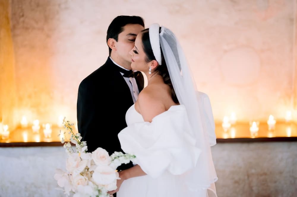 Tender close-up of the groom kissing the bride beneath a glowing candle-lined arch.