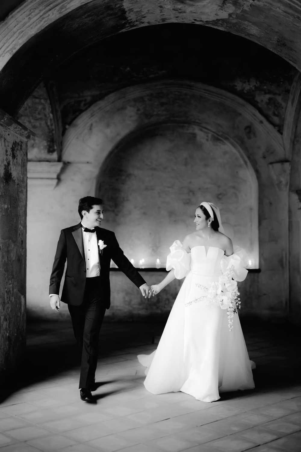 Newlyweds walk hand in hand through a vaulted stone passage — black-and-white editorial couple portrait.