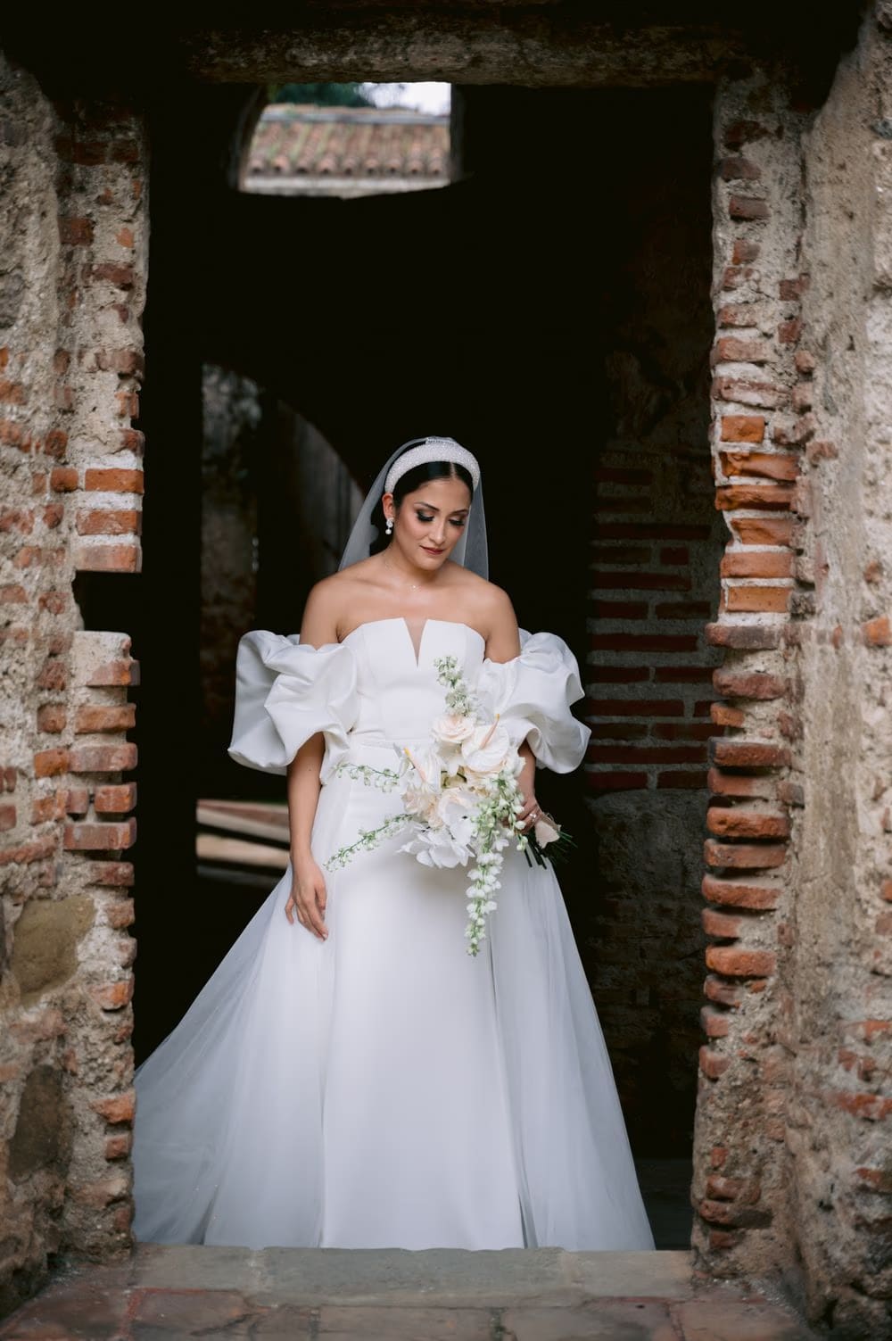 Bride framed in a rustic brick doorway, head bowed toward her cascading orchid bouquet.