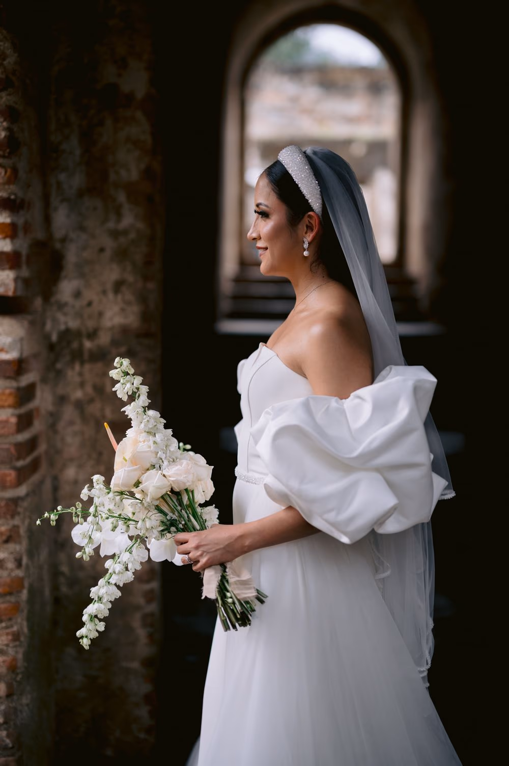 Side bridal portrait — bouquet of white orchids and roses against a softly blurred stone arch background.
