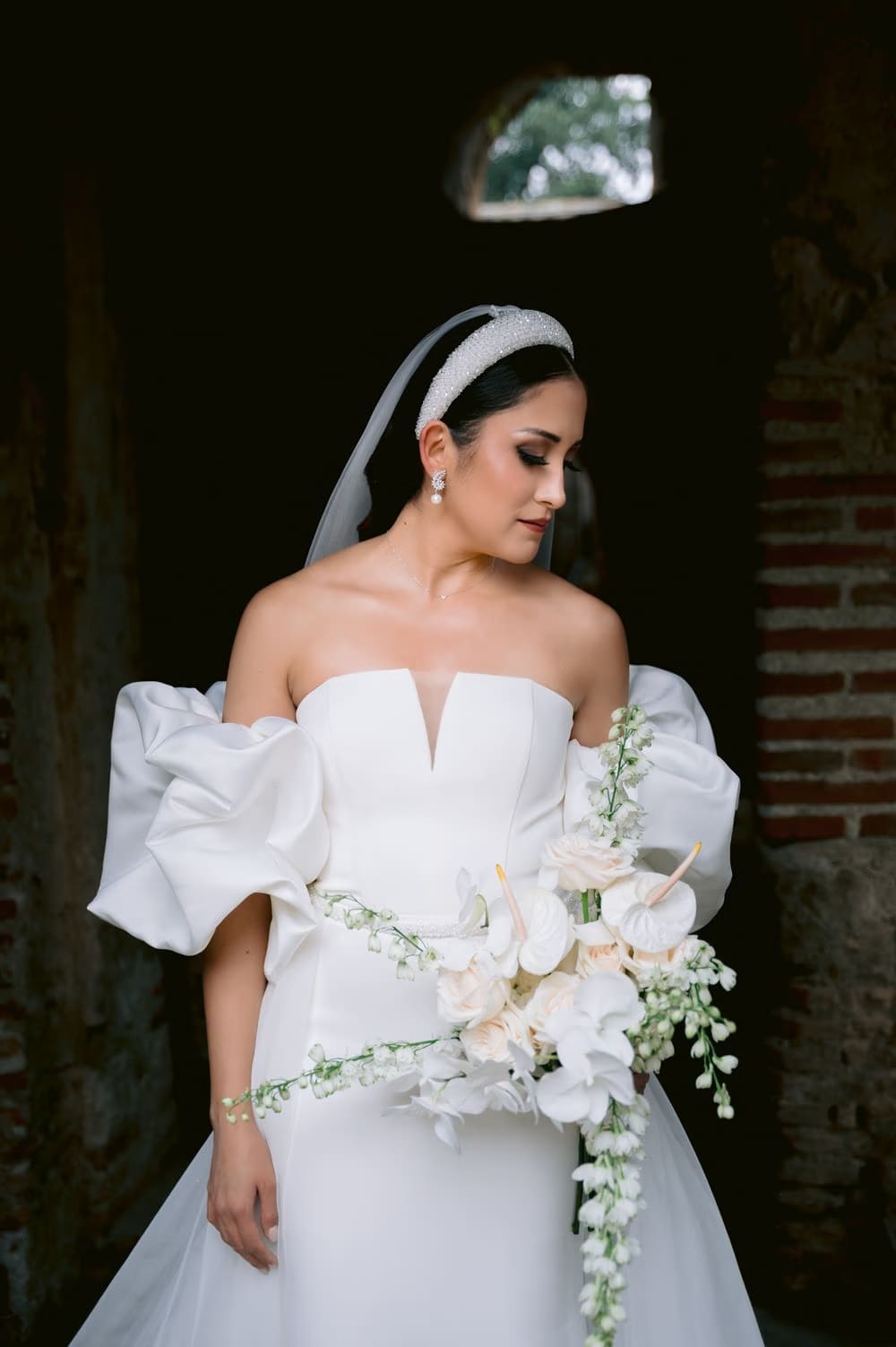 Bride looks down at her cascading bouquet in a dark stone passage — moody editorial wedding portrait.