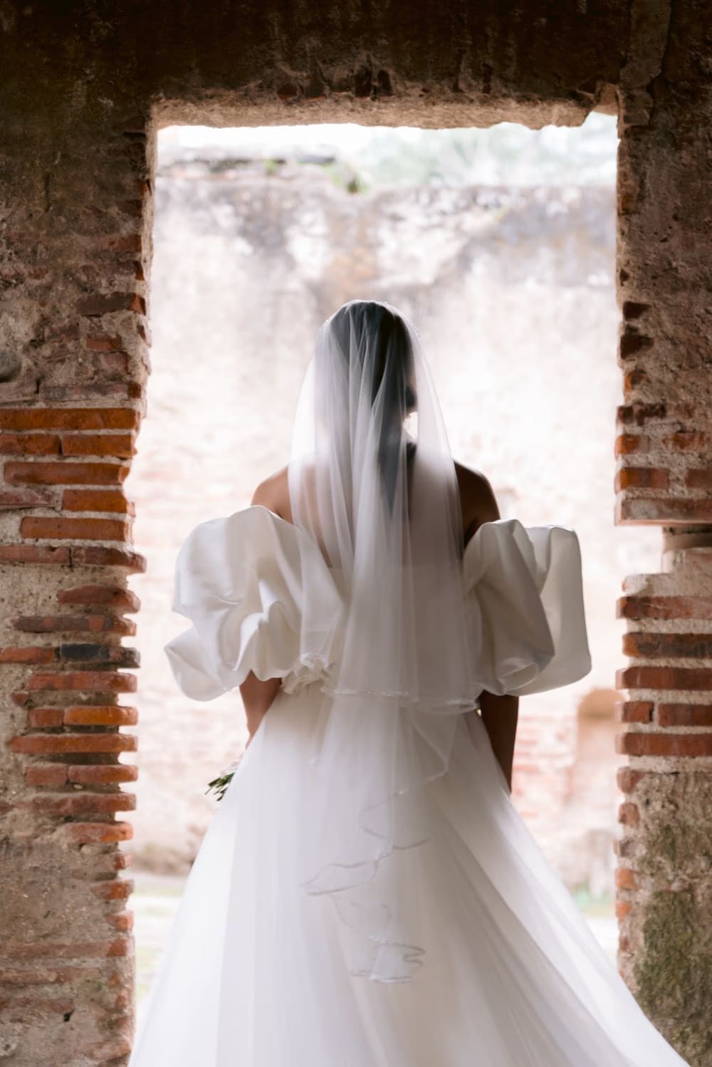 Bride photographed from behind walking through a rough brick threshold — train and veil flowing.