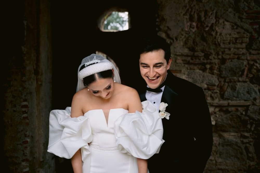 Bride dips her head laughing as the groom looks on — candid couple moment by a stone wall.