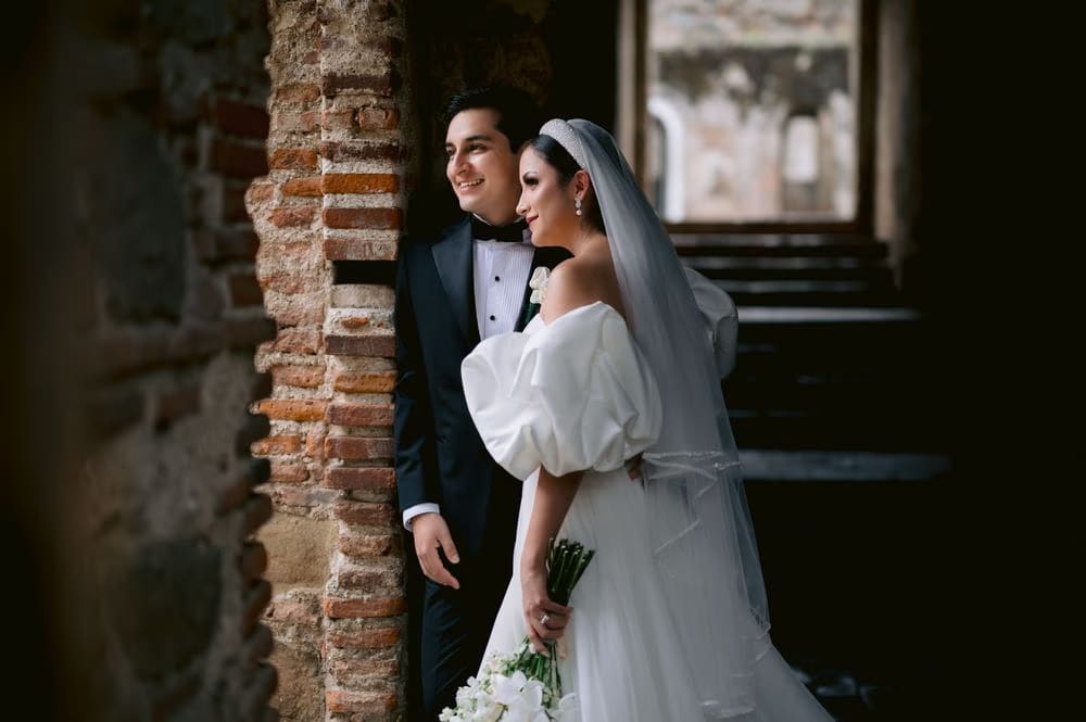 Bride and groom share a quiet smile against a brick column — candid editorial couple frame.