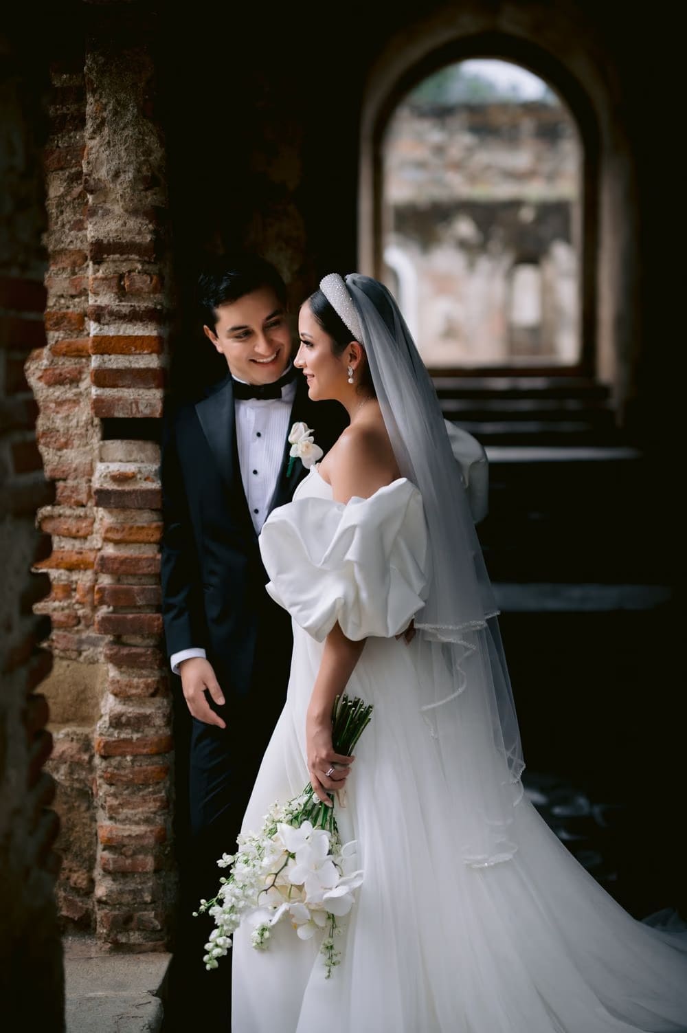 Groom smiles at his bride who looks past camera, holding her cascade bouquet — soft daylight couple portrait.