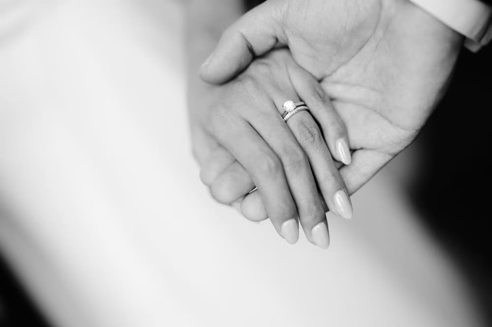 Close-up of clasped hands — bride's diamond ring rests on the groom's palm in black-and-white.