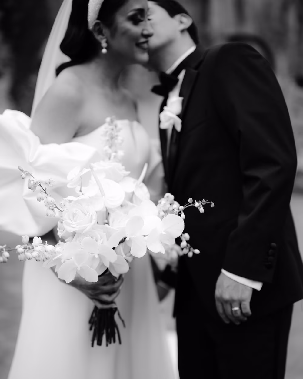 Soft-focus black-and-white close-up of the bridal bouquet of white orchids with the groom kissing the bride behind.