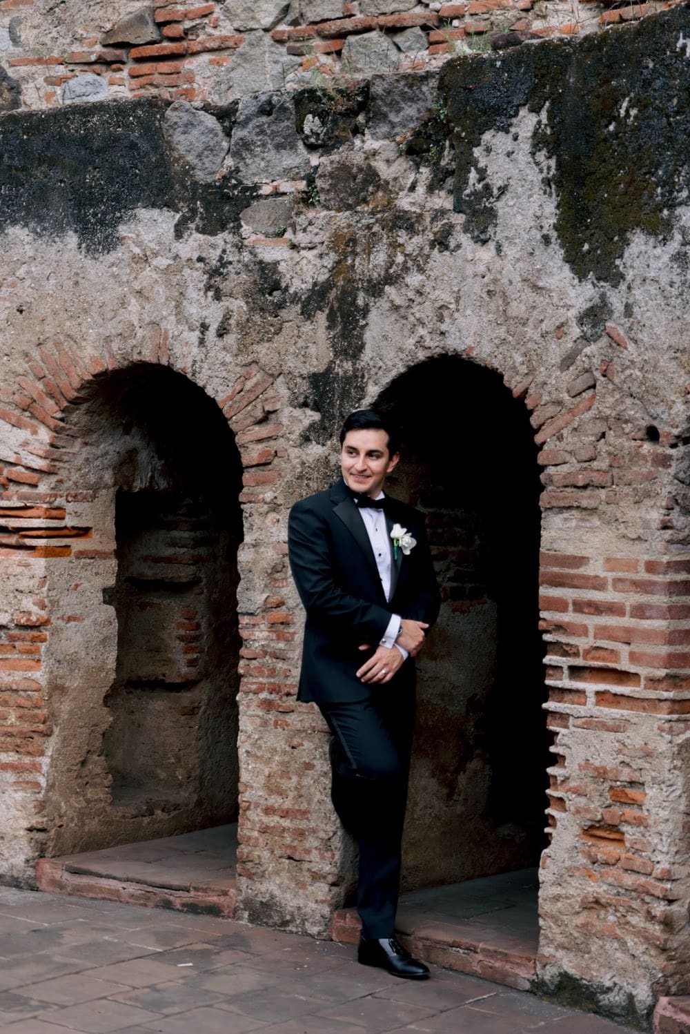 Groom Josue stands smiling between two stone niches of the convent ruins — full-length portrait.