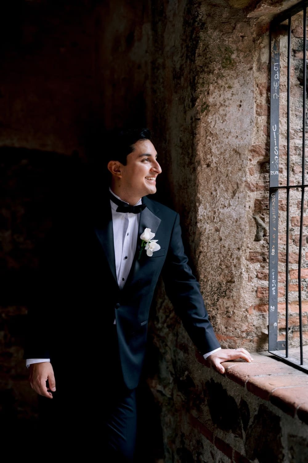 Groom looks out a barred stone window during a quiet moment — editorial black-tie portrait.