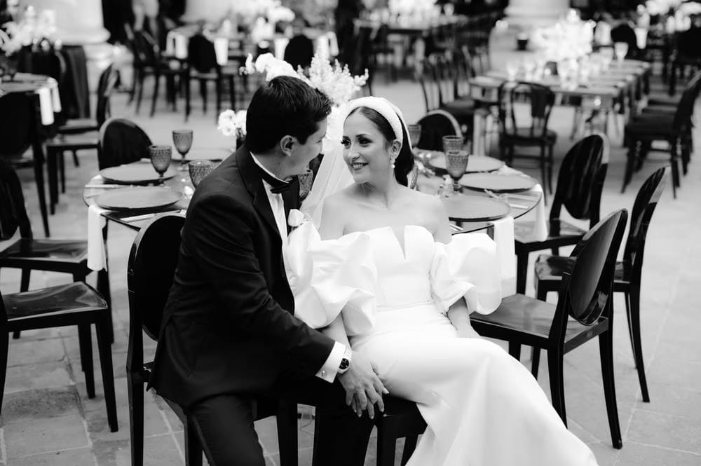 Black-and-white candid of the couple in conversation at the dinner table of the courtyard reception.