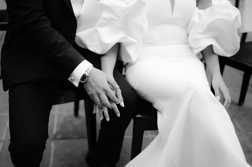 Close-up of the couple's hands gently joined on their laps — wedding ring detail in black-and-white.