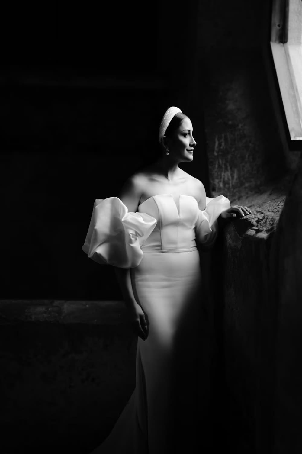 Bride glances toward a window in a dark stone room — moody black-and-white editorial portrait.