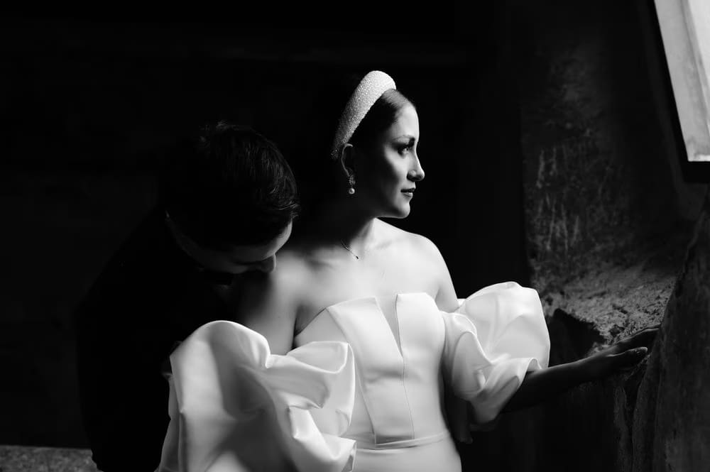Groom kisses the bride's bare shoulder as she gazes off camera — intimate black-and-white frame.