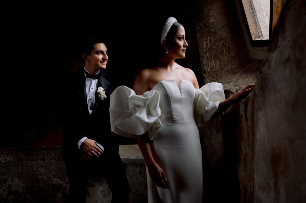 Bride leans against a stone wall while the groom sits behind — editorial low-light couple portrait.