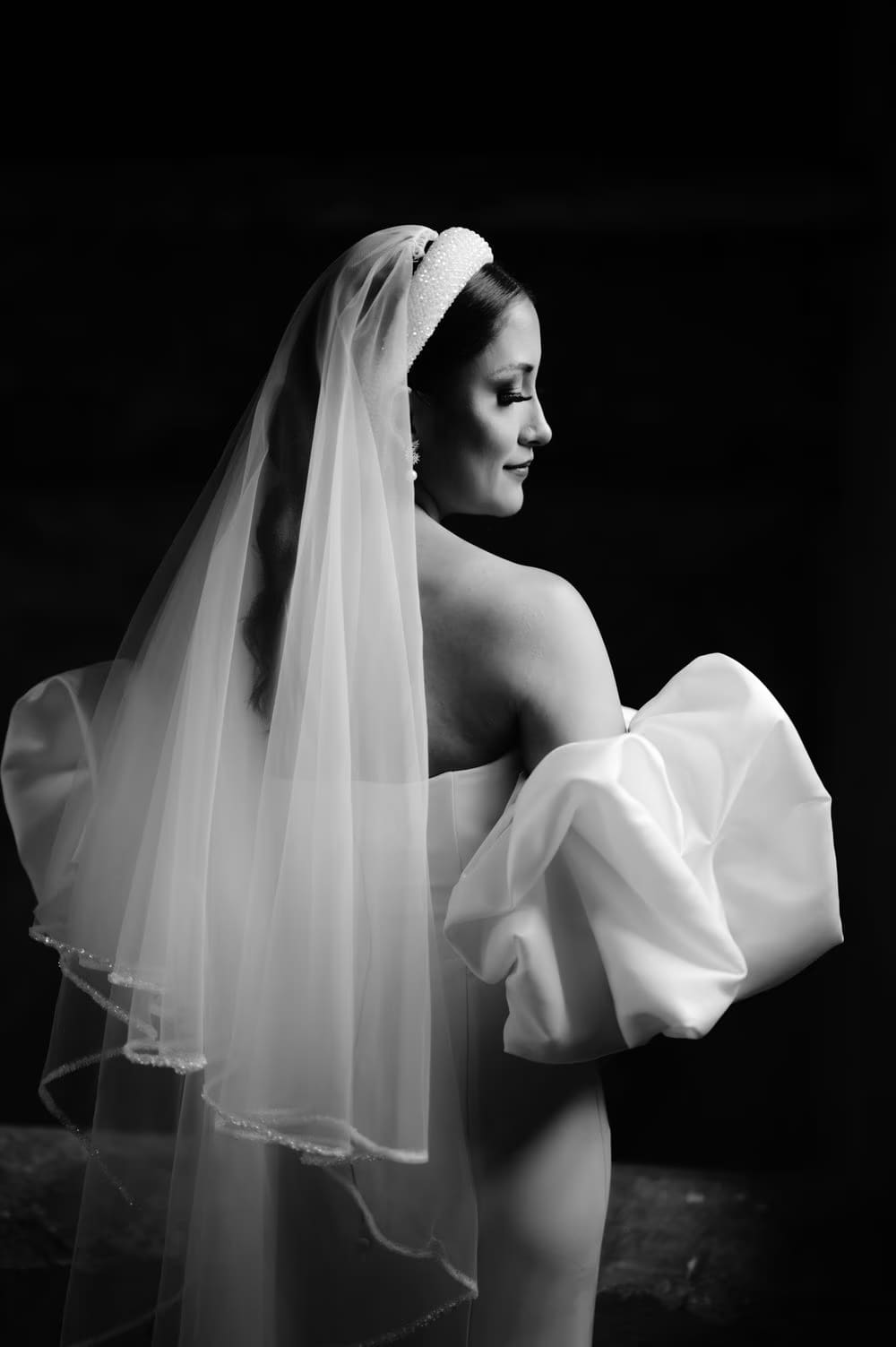 Bride photographed from behind with veil flowing down — moody studio-style black-and-white portrait.