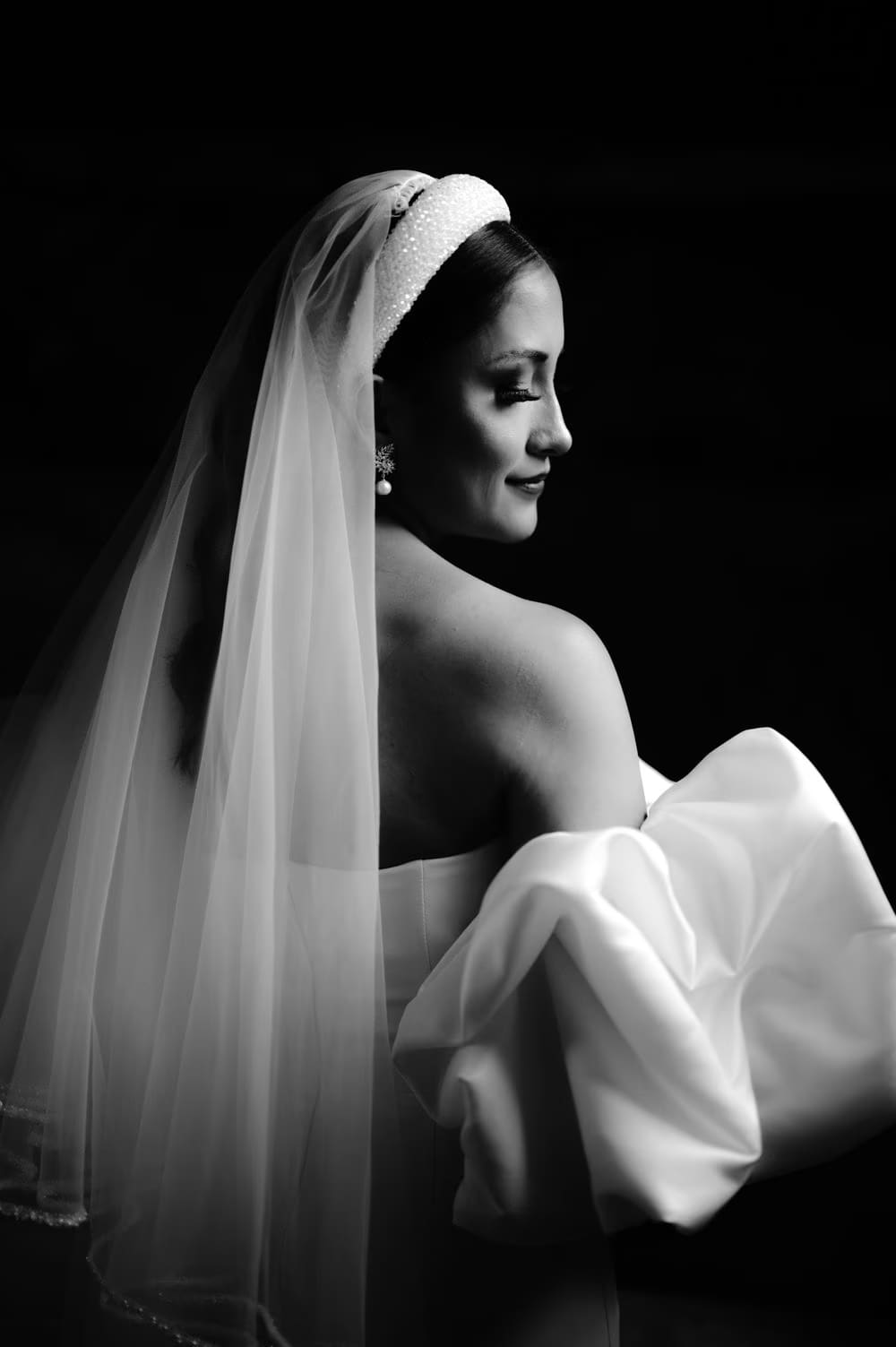 Black-and-white bridal portrait — bride in profile with crystal headband, veil and pearl drop earring against dark backdrop.