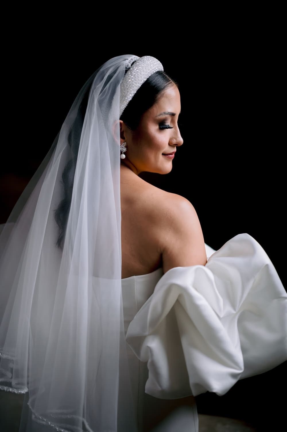 Bride glances over her bare shoulder with smoky eye makeup and cathedral veil cascading behind.