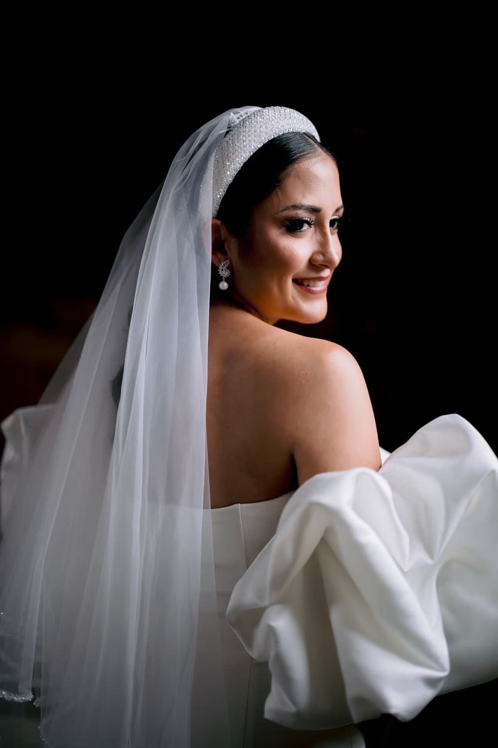 Bride smiles softly looking back — close editorial portrait highlighting headband and pearl drop earring.