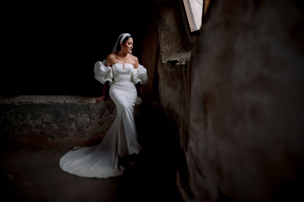 Full-length bride leaning against a stone wall in mermaid silhouette — black-and-white editorial frame.
