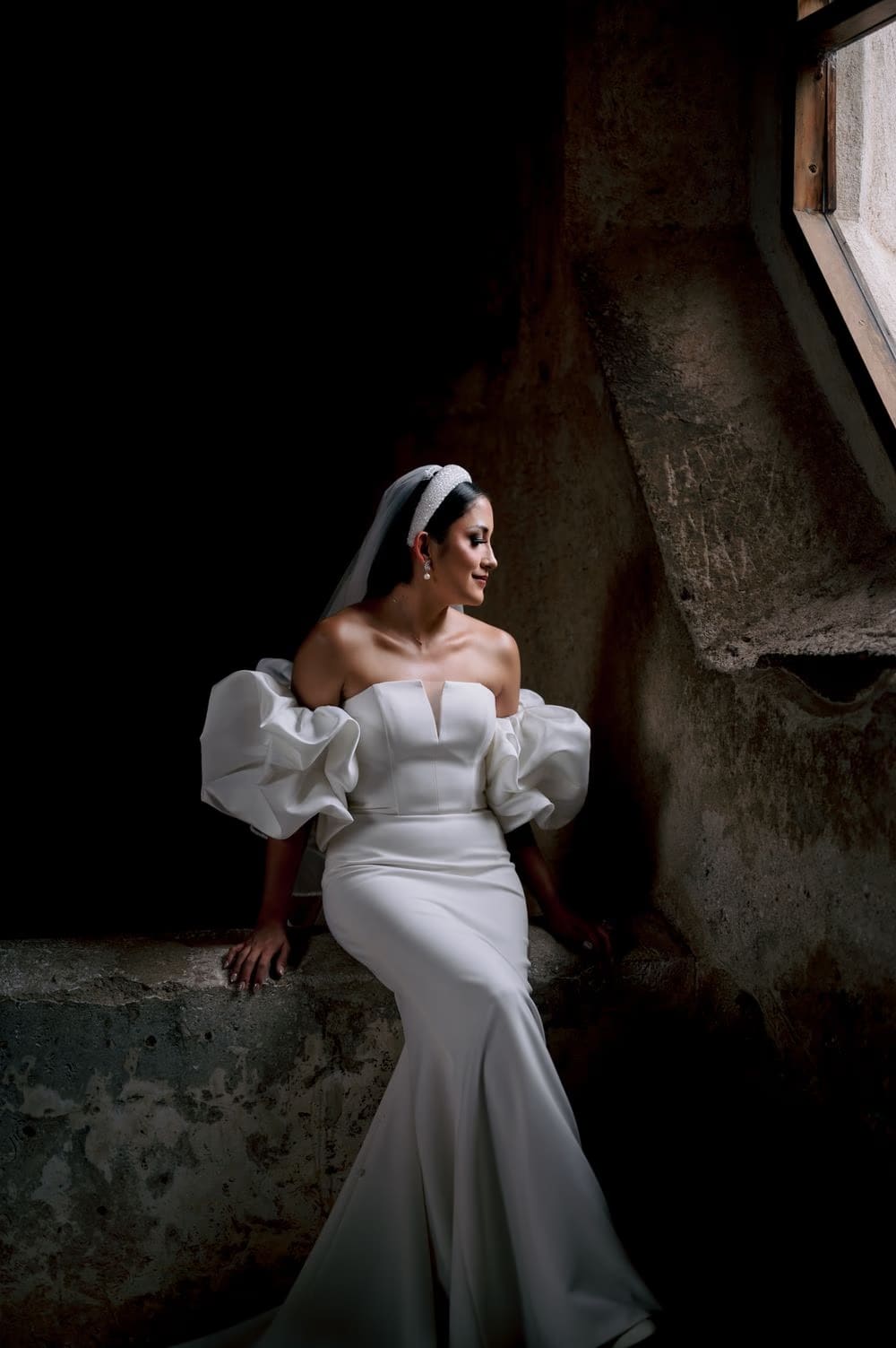 Bride seated on a stone ledge facing a tall window — soft natural light from above.