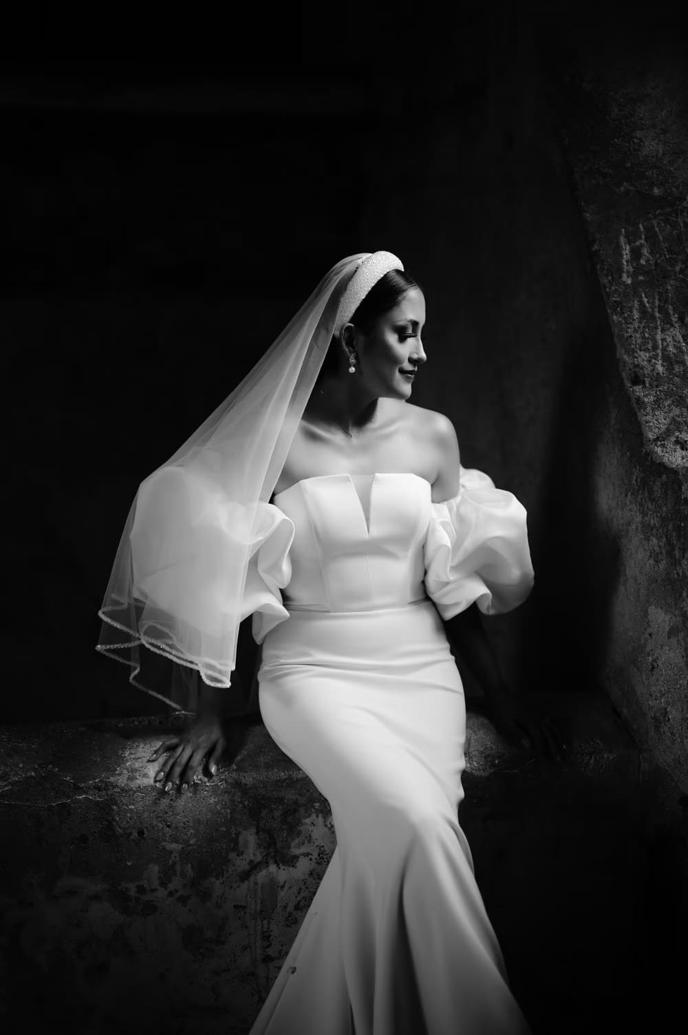 Bride sits on a stone ledge in mermaid silhouette with veil over one side — black-and-white editorial.