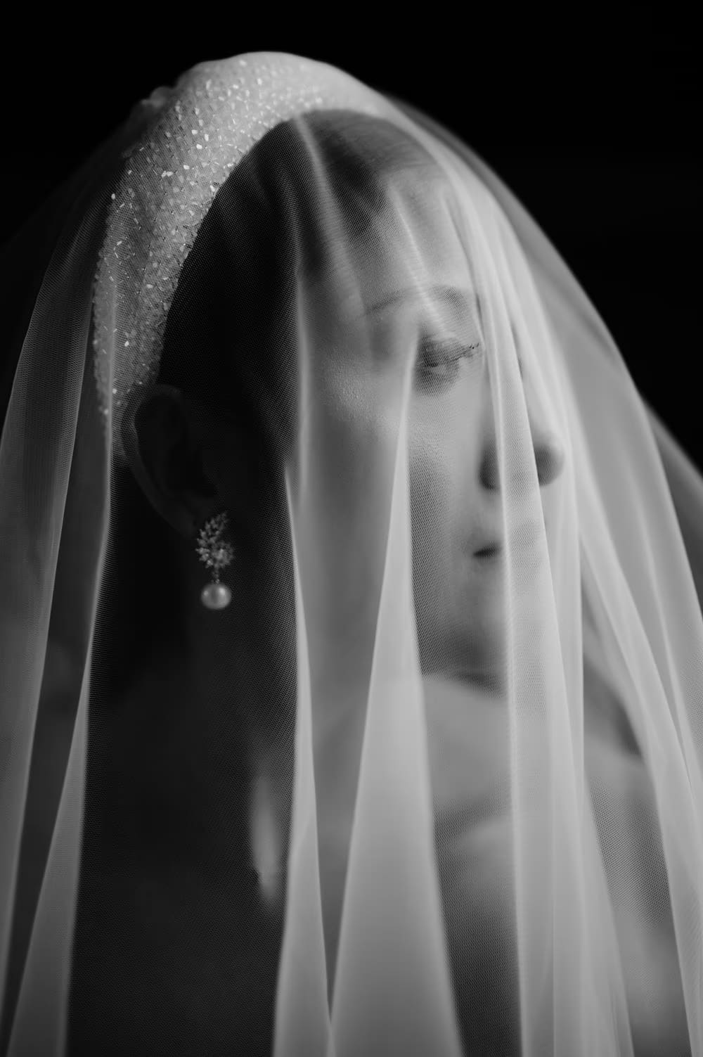 Bride veiled in tulle with pearl earring catching the light — ethereal black-and-white close portrait.