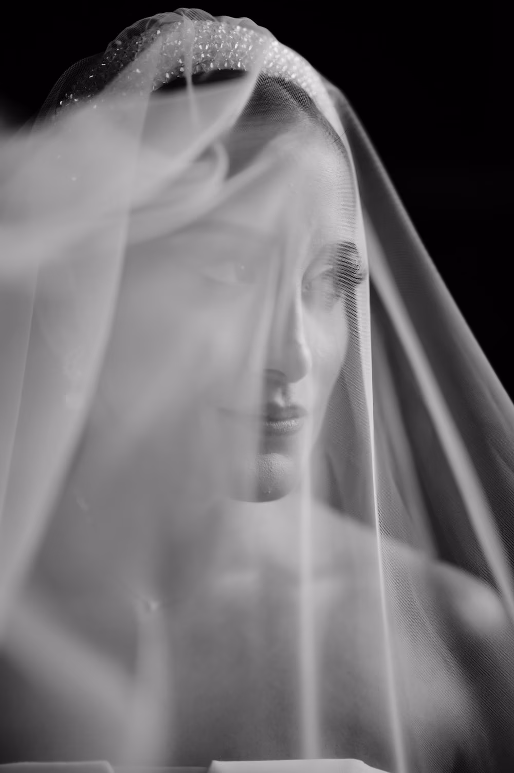 Bride's face seen through layered tulle veil — soft, poetic black-and-white close-up.