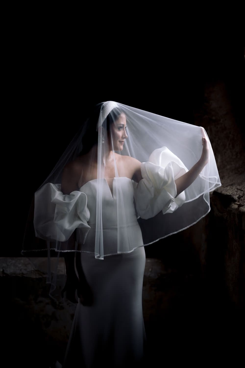 Bride lifts her veil with both hands creating a glowing canopy — editorial black-and-white frame.