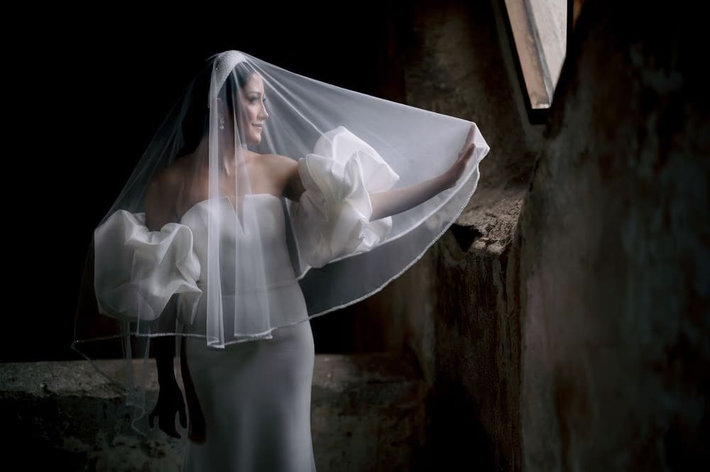 Bride lifts the veil and turns toward window light against rough stone walls — fine-art portrait.