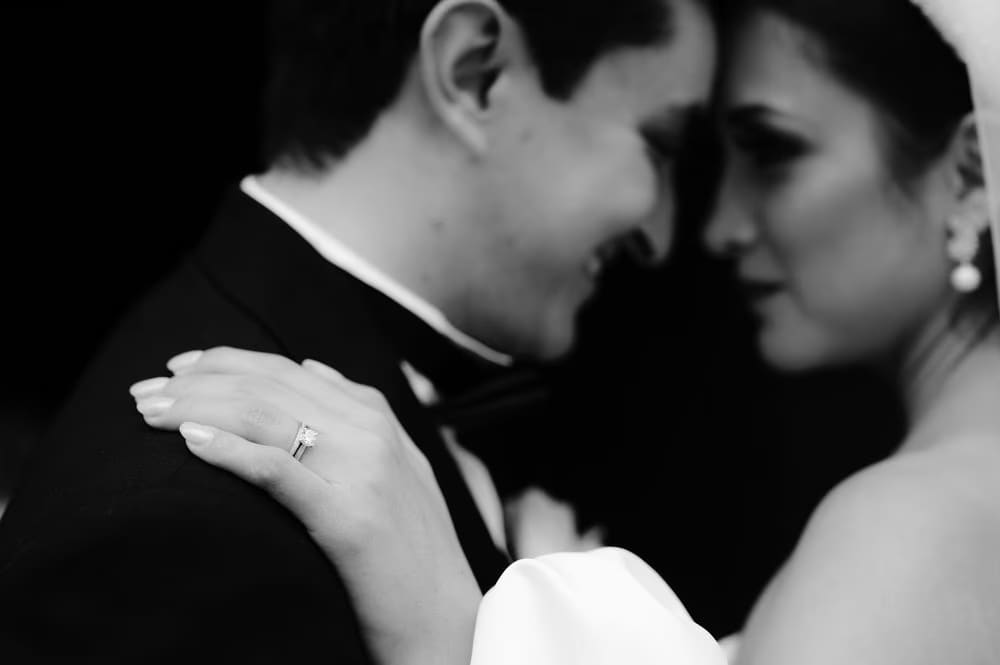 Close-up of bride's hand on the groom's lapel revealing the diamond wedding ring — black-and-white detail.