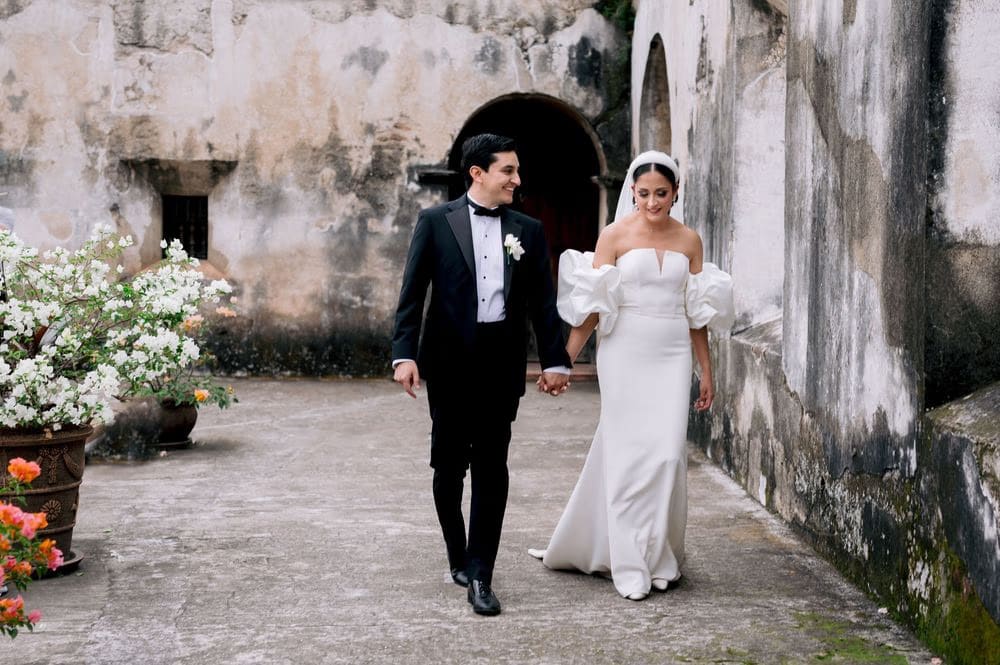Newlyweds stroll hand in hand through a colonial courtyard with bougainvillea — destination wedding portrait.