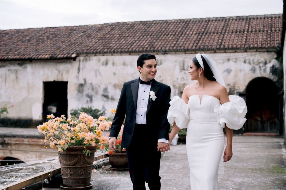 Bride and groom hold hands strolling past terracotta planters with bougainvillea on a tile rooftop.