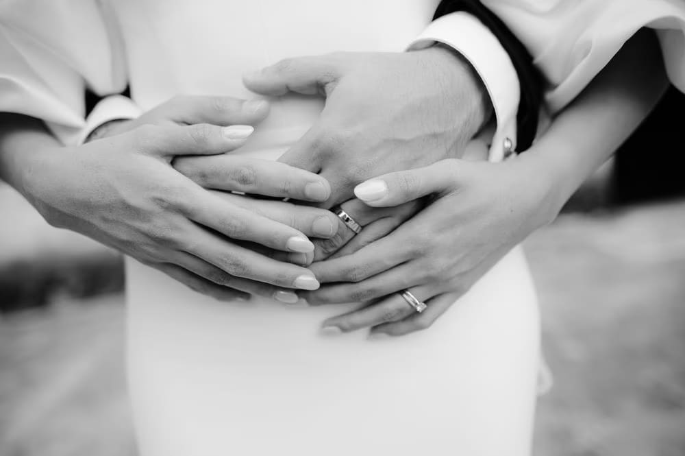 Close-up of the couple's hands intertwined around the bride's waist — both wedding bands visible.