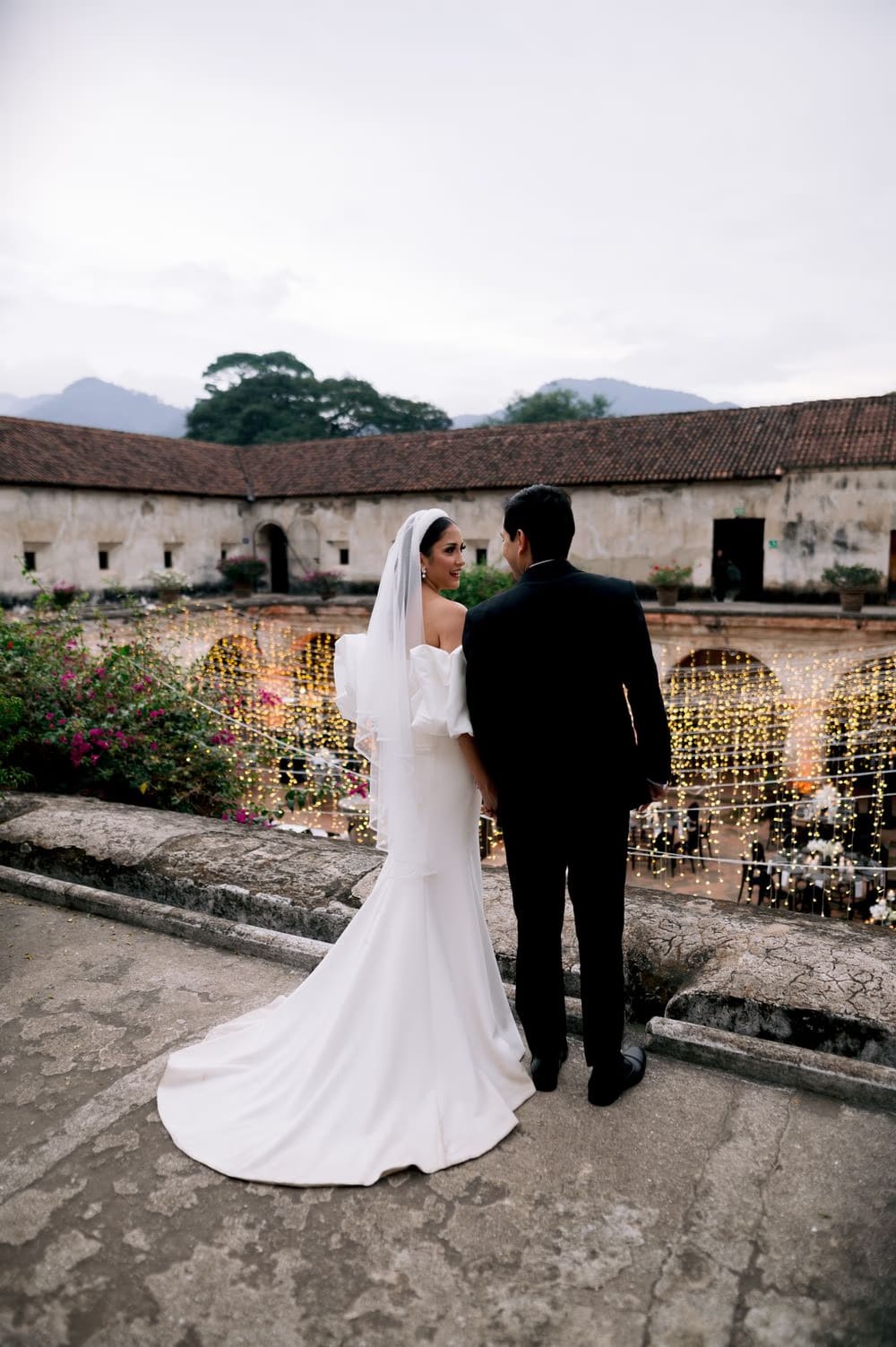 Bride and groom seen from behind on a terrace overlooking the string-lit reception courtyard at dusk.