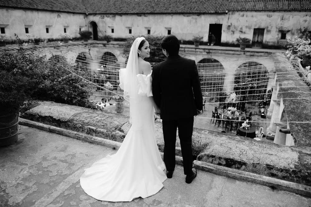 Bride and groom look out at the lit-up courtyard reception from a terrace above — golden glow below.