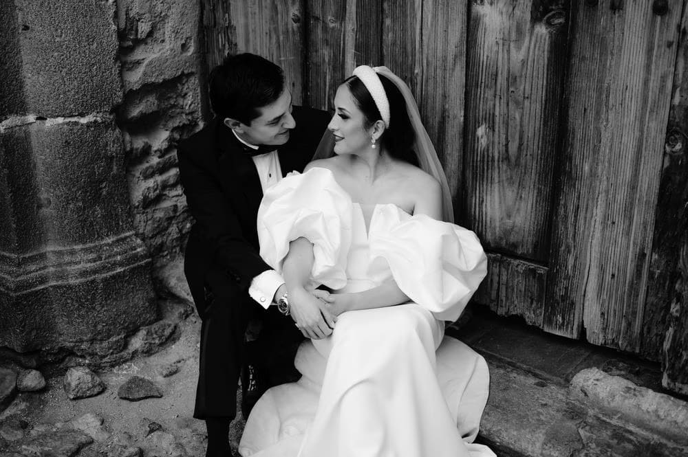 Newlyweds laugh together seated against a worn wooden door — candid black-and-white couple frame.