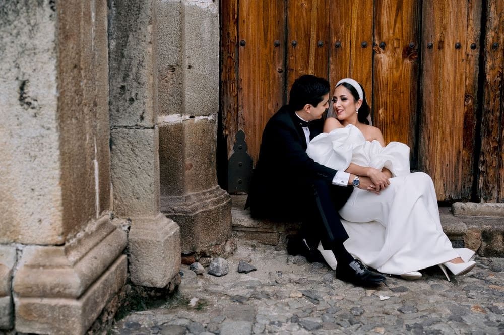 Bride and groom sit on cobblestones beside an old wooden door, holding each other in intimate quiet.