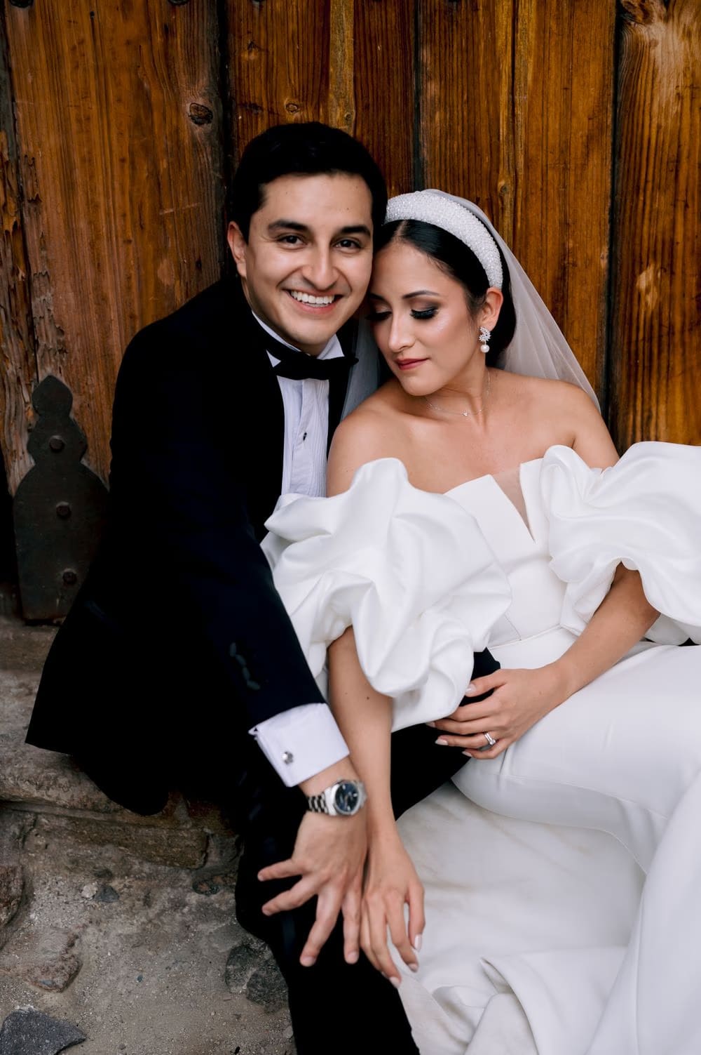 Bride leans on the groom's shoulder against a wooden colonial door — relaxed editorial couple portrait.