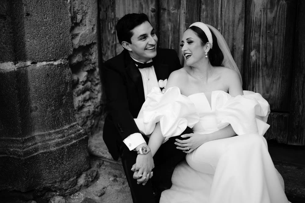 Bride and groom share an open-mouthed laugh seated by an old door — joyful black-and-white candid.