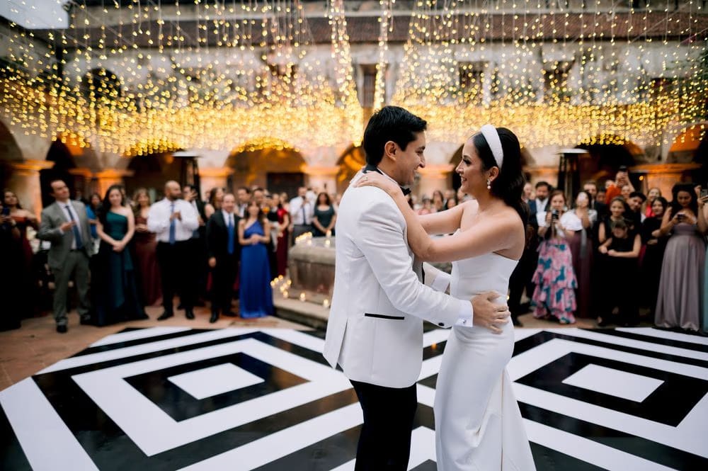 Bride and groom share their first dance under a canopy of string lights in the cloister courtyard — guests watch.