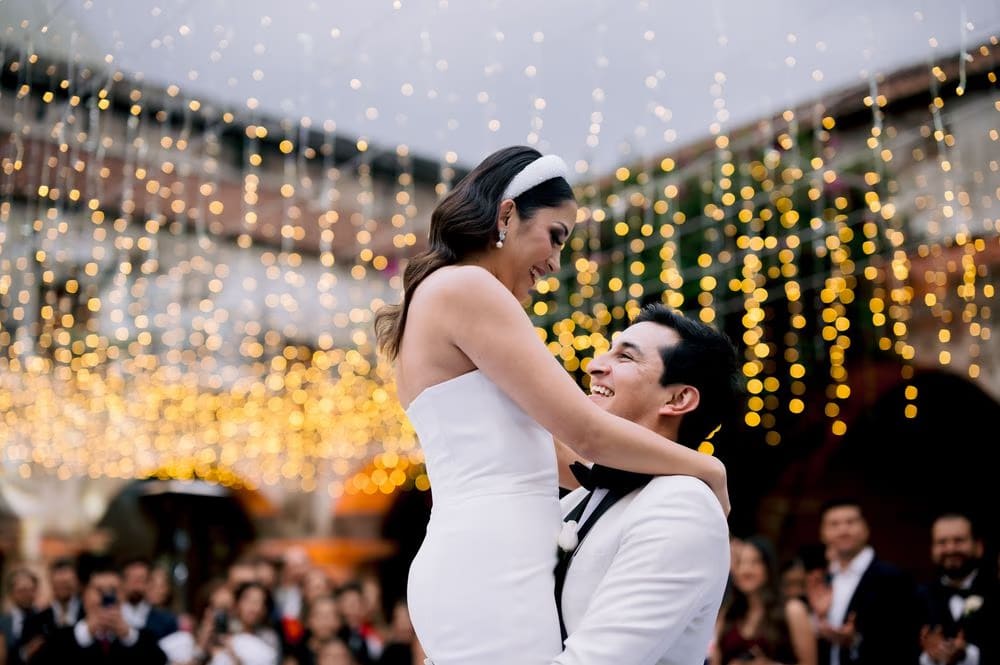 Groom lifts the bride in his arms on the dance floor as she laughs — fairy-light reception backdrop.