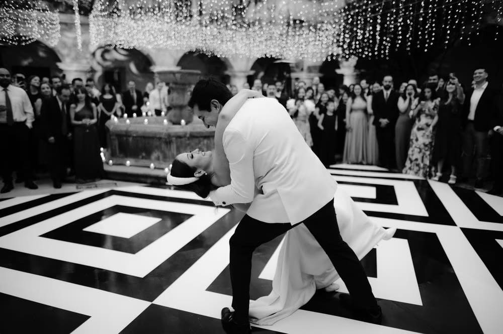 Dramatic dance-floor dip — groom in white tuxedo dips the bride on the geometric floor as guests cheer.