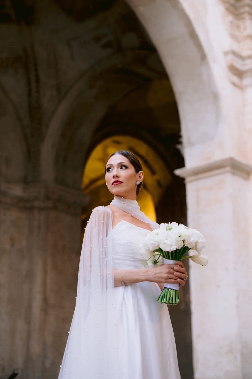 Bride Andrea holds a white ranunculus bouquet beneath a carved stone archway at Capuchinas Ruins — intimate destination wedding in Antigua Guatemala.