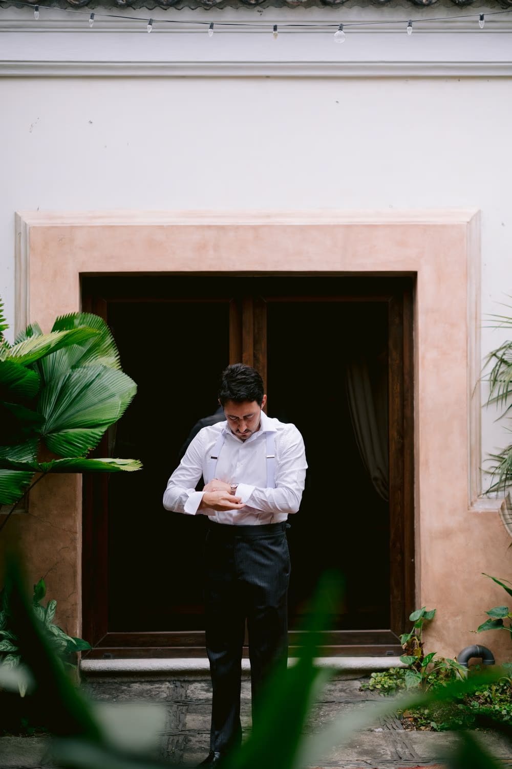 Groom Juan adjusts his cufflinks framed by a courtyard doorway and tropical leaves during getting-ready at La Antigua wedding morning.