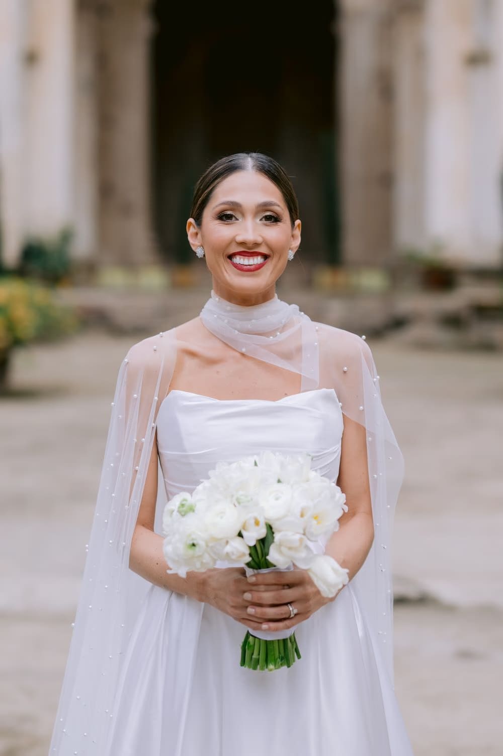 Bride smiles holding her white bouquet, satin gown and pearl-dotted cape catching soft daylight at Capuchinas Ruins, Antigua Guatemala.