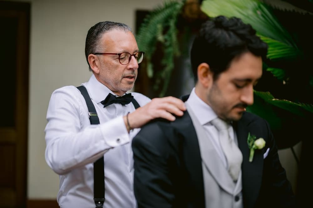 Father in suspenders helps the groom into his tuxedo jacket beside tropical foliage — family wedding morning in Antigua.