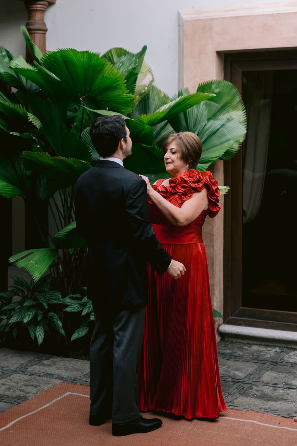 Mother of the groom in a red ruffled gown meets her son in a leafy courtyard before the ceremony in Antigua Guatemala.
